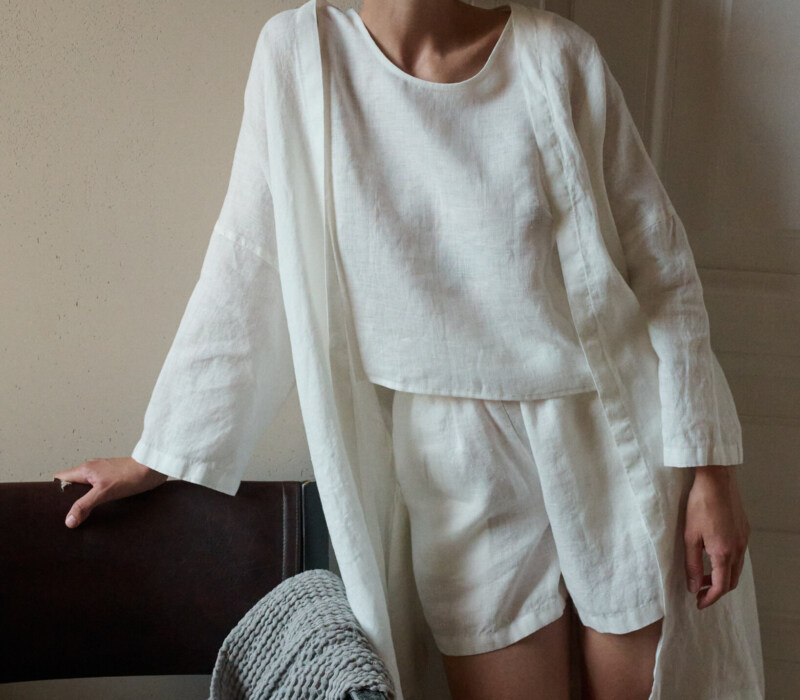 Woman wearing an all-white lightweight linen outfit consisting of a loose-fitting sleeveless top, shorts, and an short open robe