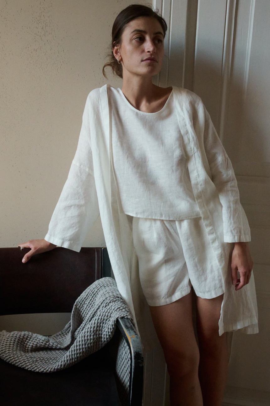 Woman wearing an all-white lightweight linen outfit consisting of a loose-fitting sleeveless top, shorts, and an short open robe