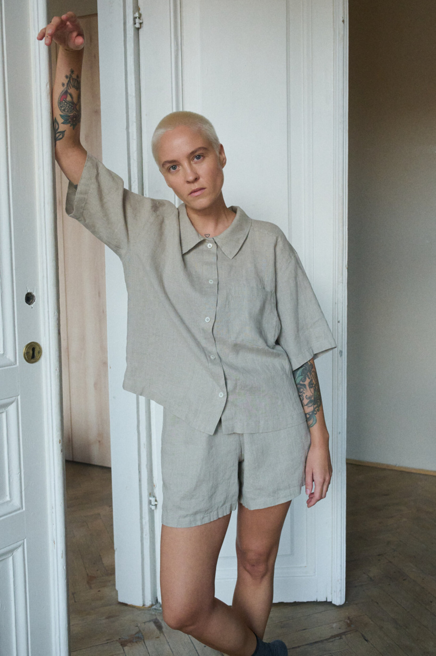 Model wearing a relaxed, natural gray linen two-piece pajama set, including a loose-fitting, short-sleeve button-up shirt with a wide collar and a straight hem that falls just above the waist, and matching shorts in a simple, casual design, reaching mid-thigh