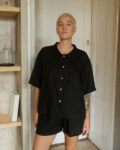 Woman wearing a relaxed black linen two-piece set, featuring a loose short-sleeve button-up shirt and mid-thigh shorts, offering a minimalist and casual look