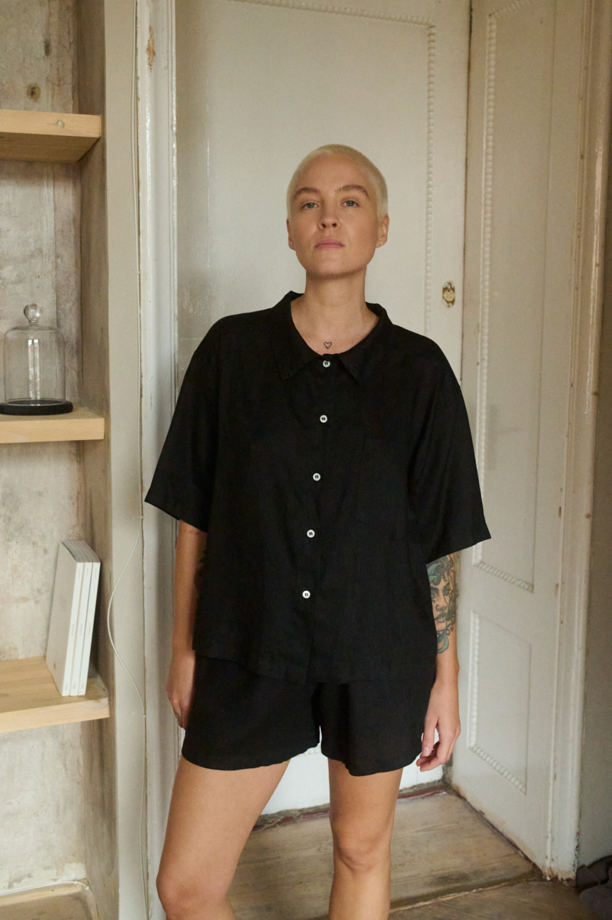 Woman wearing a relaxed black linen two-piece set, featuring a loose short-sleeve button-up shirt and mid-thigh shorts, offering a minimalist and casual look