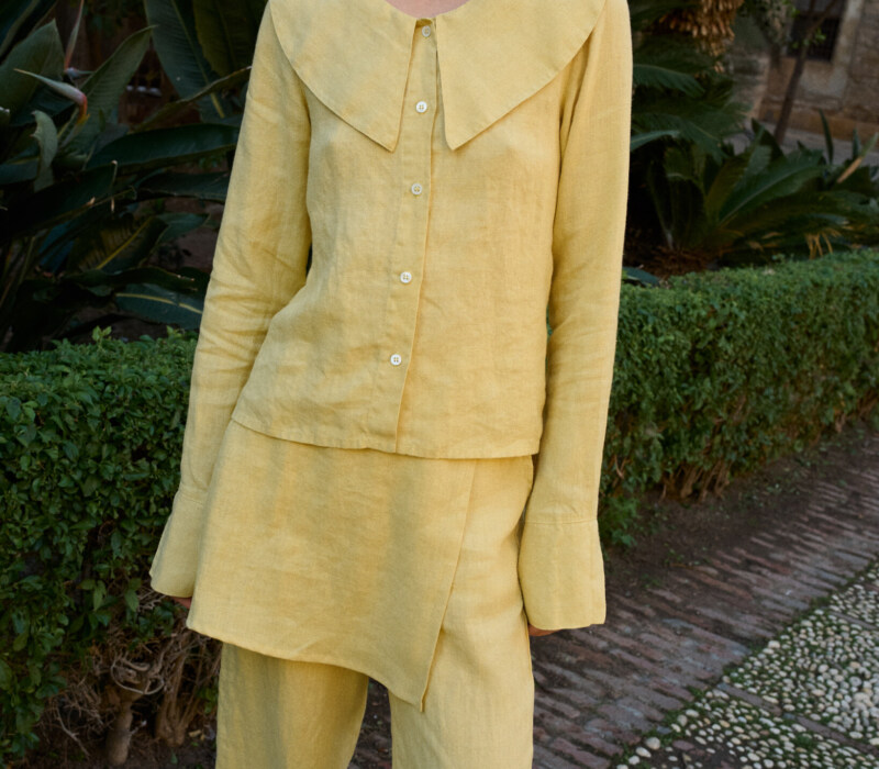 Model wearing a butter-yellow linen three-piece set, including a tailored button-down blouse with a bold oversized collar, an asymmetrical layered skirt panel, and relaxed wide-leg trousers