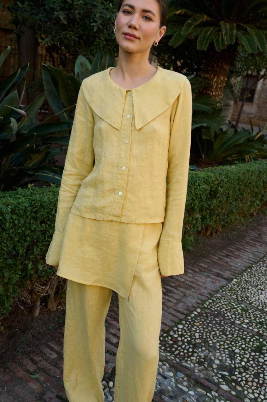 Model wearing a butter-yellow linen three-piece set, including a tailored button-down blouse with a bold oversized collar, an asymmetrical layered skirt panel, and relaxed wide-leg trousers