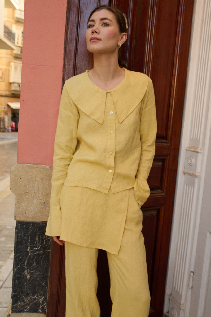 Model showcasing a butter-yellow linen three-piece set, including a shirt with a striking oversized collar, an asymmetrical layered skirt, and relaxed wide-leg trousers