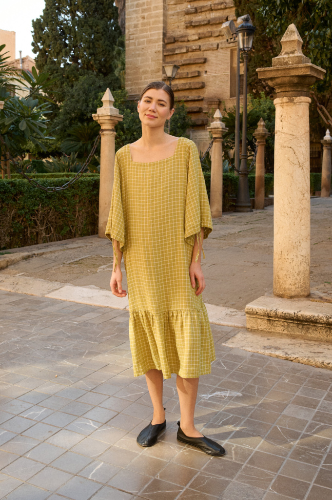 Model wearing a soft pickle-green linen dress with subtle white grid checks, featuring a square neckline, wide three-quarter length sleeves, and a gathered ruffle hem that falls to mid-calf