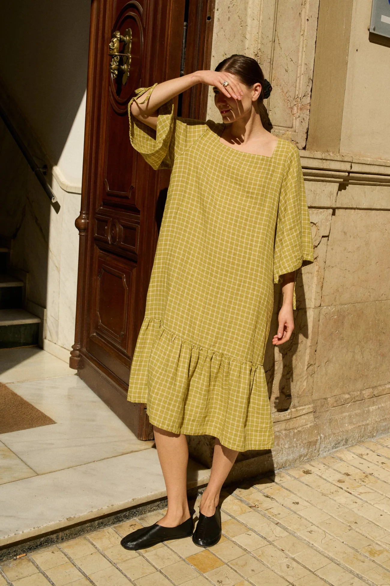 Model in a soft pickle-green linen dress with white grid checks, showcasing a square neckline, wide three-quarter length sleeves, and a flowing, gathered ruffle hem that falls to mid-calf, offering a relaxed look