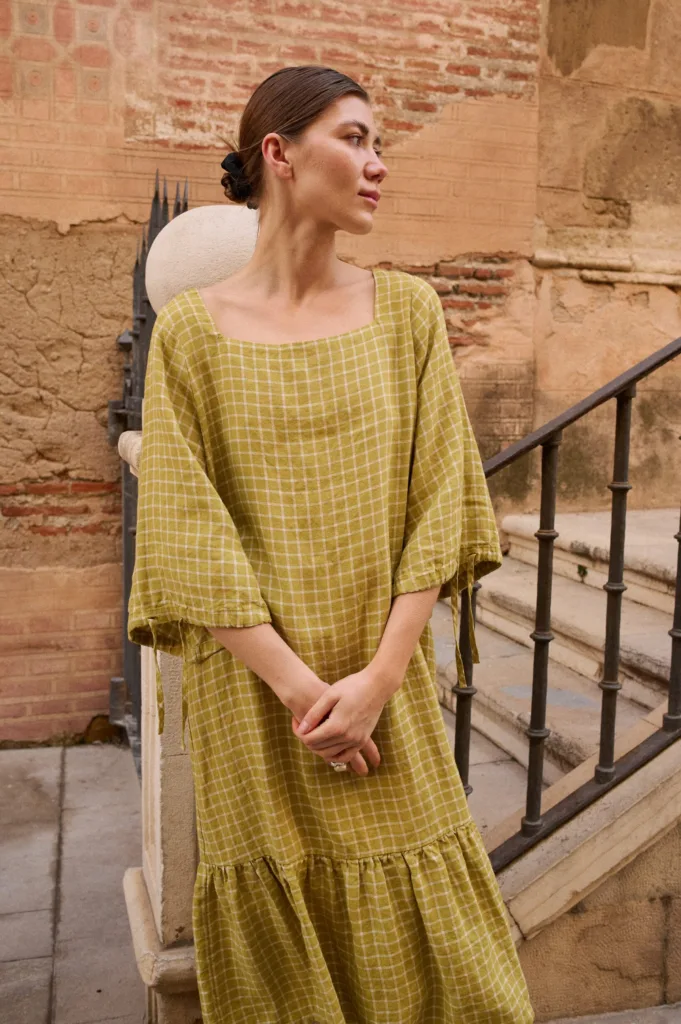 Model wearing a pickle-green and white checked linen dress with a wide square neckline, three-quarter length sleeves gathered at the cuffs, a dropped waistline, and a voluminous ruffle hem