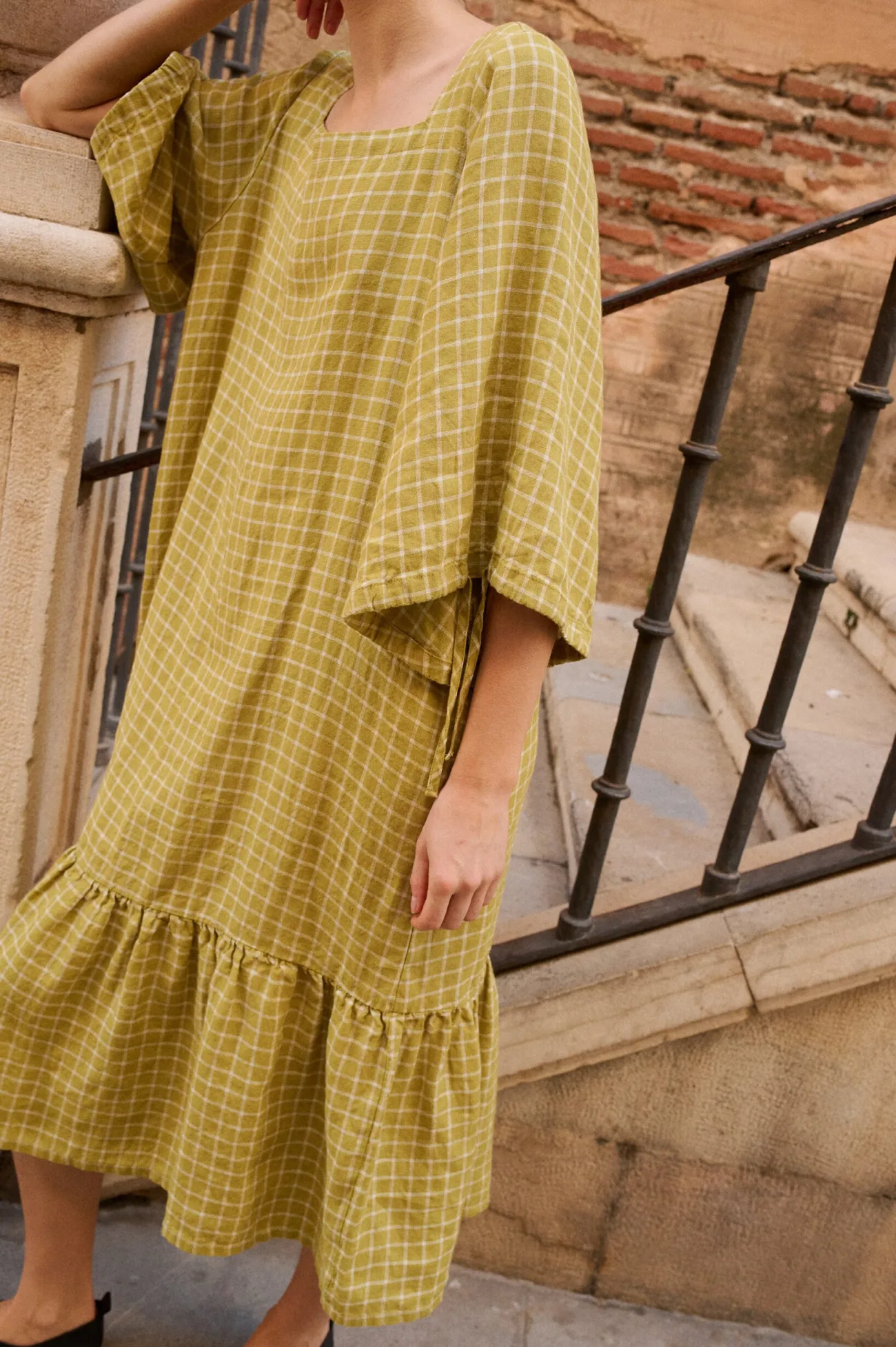 Model wearing a soft pickle checks linen dress featuring a wide square neckline, three-quarter length sleeves with gathered cuffs, and a dropped waistline with a full ruffle hem that gently falls below the knee, offering a relaxed silhouette