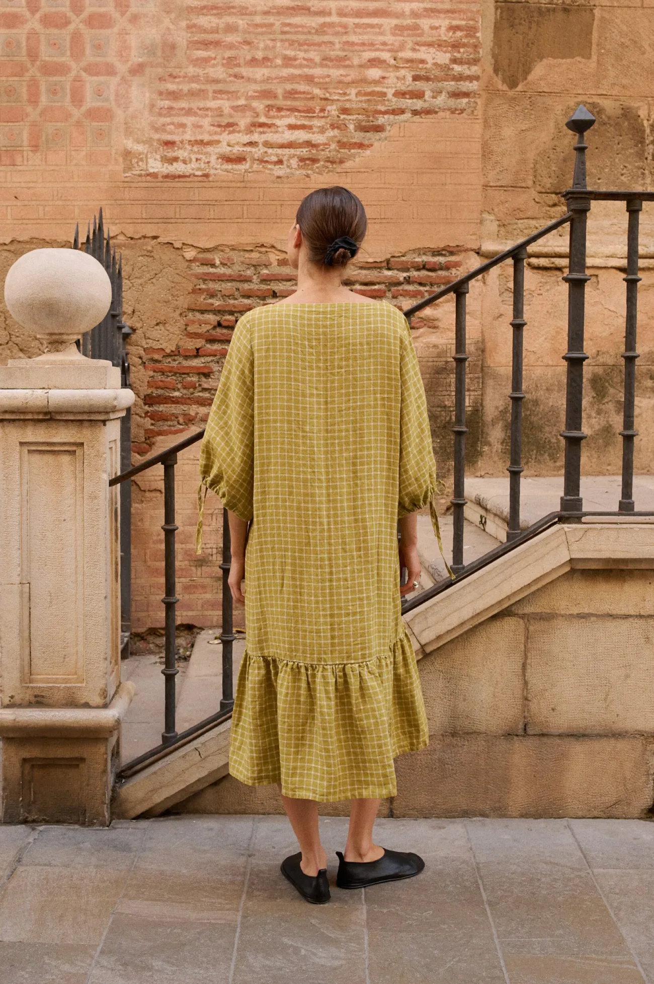 Model seen from the back, wearing a pickle-green and white checked linen dress with wide, three-quarter length sleeves and a dropped waistline, highlighted by a voluminous ruffle hem that falls below the knee