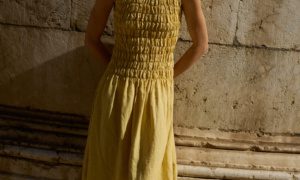 Model in a sleeveless butter-yellow linen dress with a smocked bodice that gracefully flows into a midi-length skirt, creating a light, breezy fit perfect for warm days