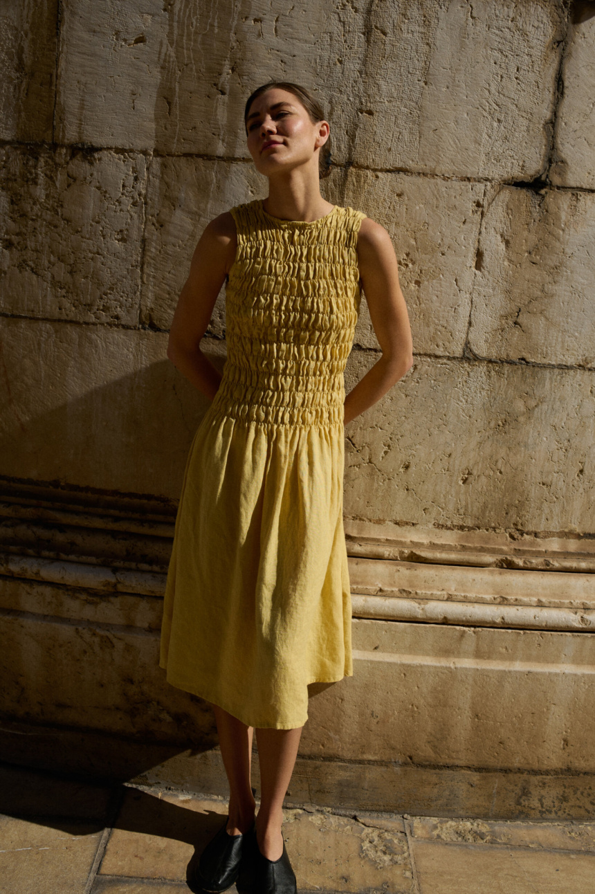 Model in a sleeveless butter-yellow linen dress with a smocked bodice that gracefully flows into a midi-length skirt, creating a light, breezy fit perfect for warm days