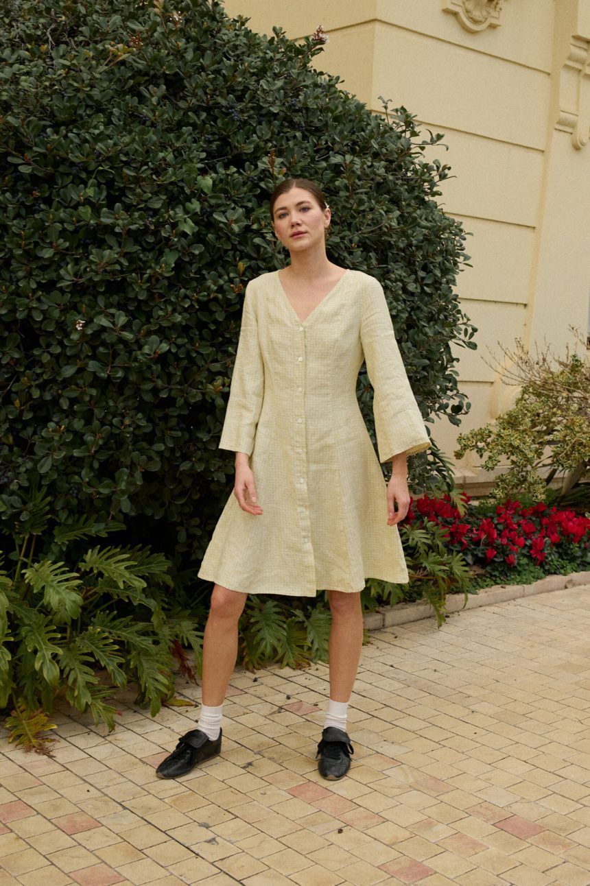 Model wearing a knee-length pale apple check dress with a V-neckline, wide three-quarter sleeves, a button-down front, and a fitted bodice that flares into an A-line silhouette