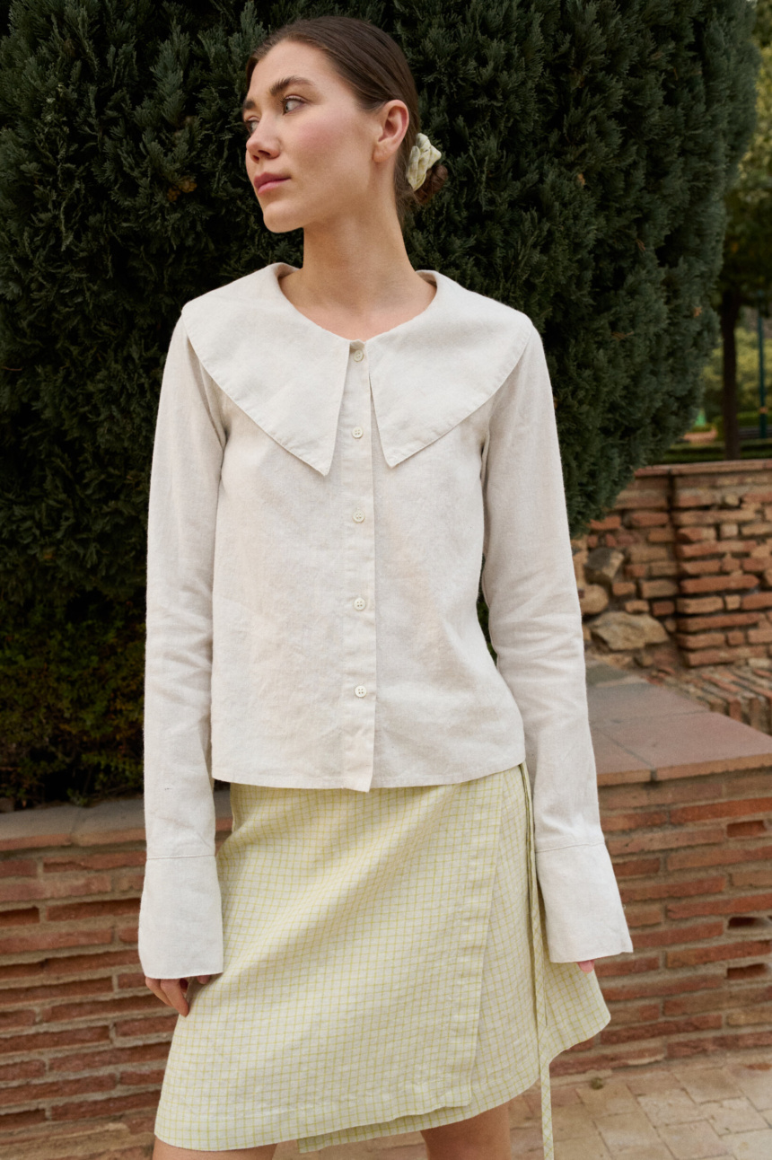 Model wearing a white cotton blend blouse with an oversized Peter Pan collar, button-down front, and full-length sleeves with subtle cuffs, paired with a pale apple checks linen wrap skirt featuring side tie closures and a subtle A-line shape