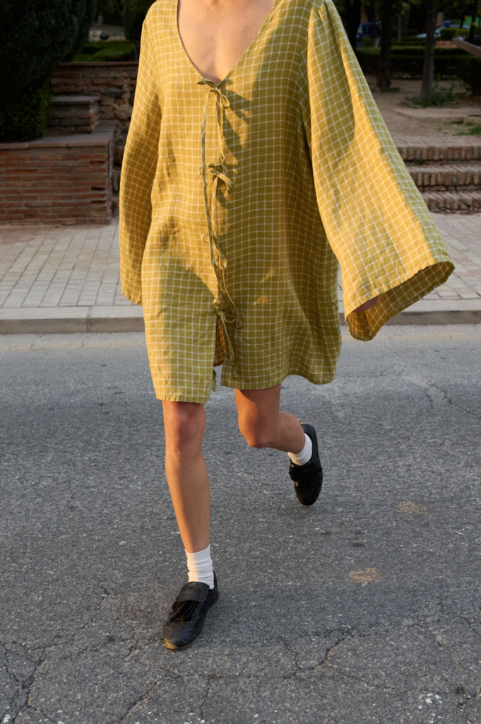 Model wearing a pickle checks linen dress with a deep V-neckline, decorative tie closures down the front, wide kimono-style sleeves, and a relaxed fit that falls to mid-thigh