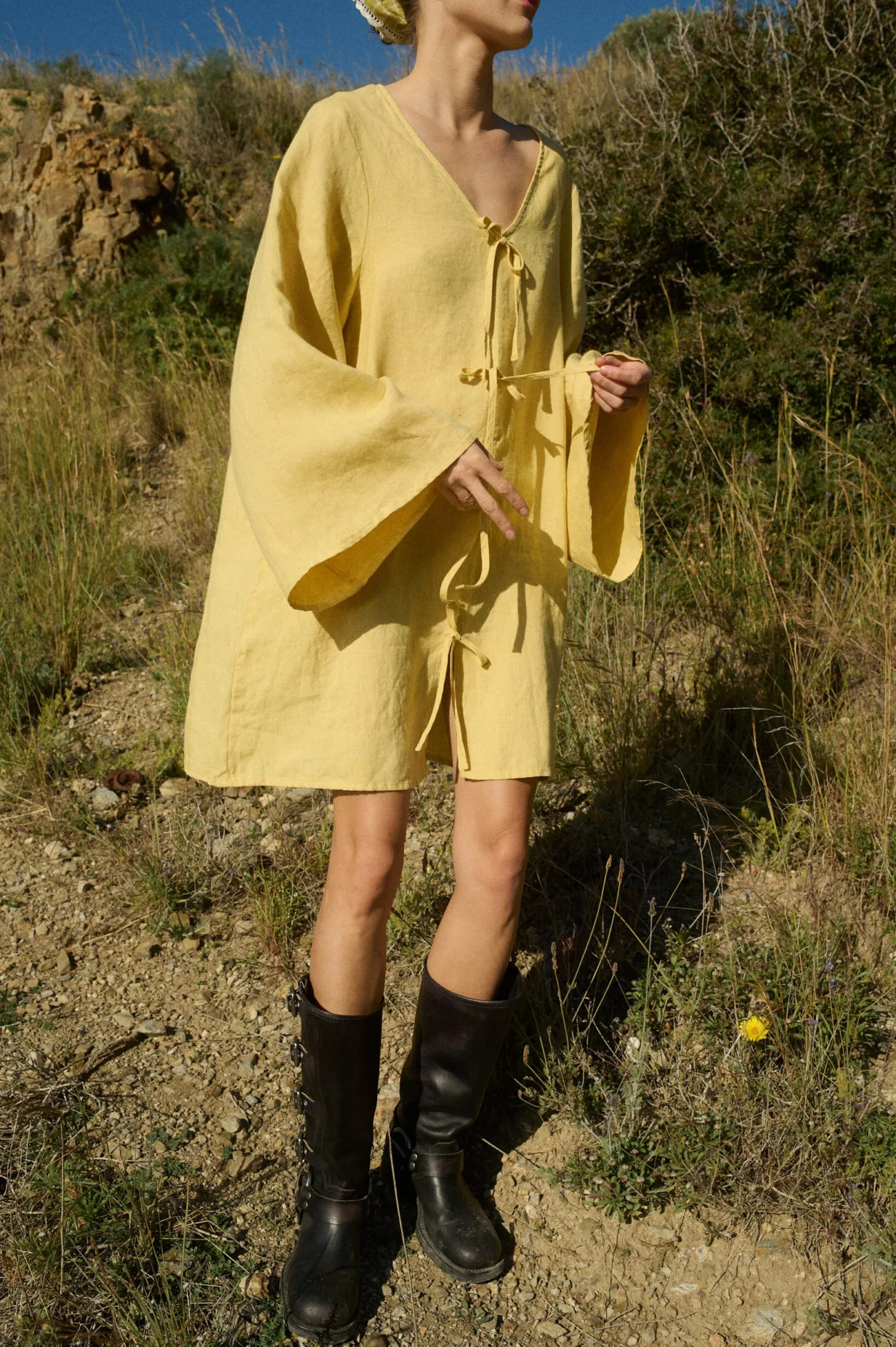 Model wearing a butter-colored mini linen dress with a deep V-neckline, four decorative front tie-bows, wide, flowy sleeves, in an oversized A-line fit