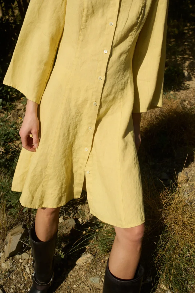 Model wearing a yellow linen dress with a V-neckline, full-length button-down front, ¾ flared sleeves, and a relaxed A-line fit, featuring vertical linen panels and godet inserts