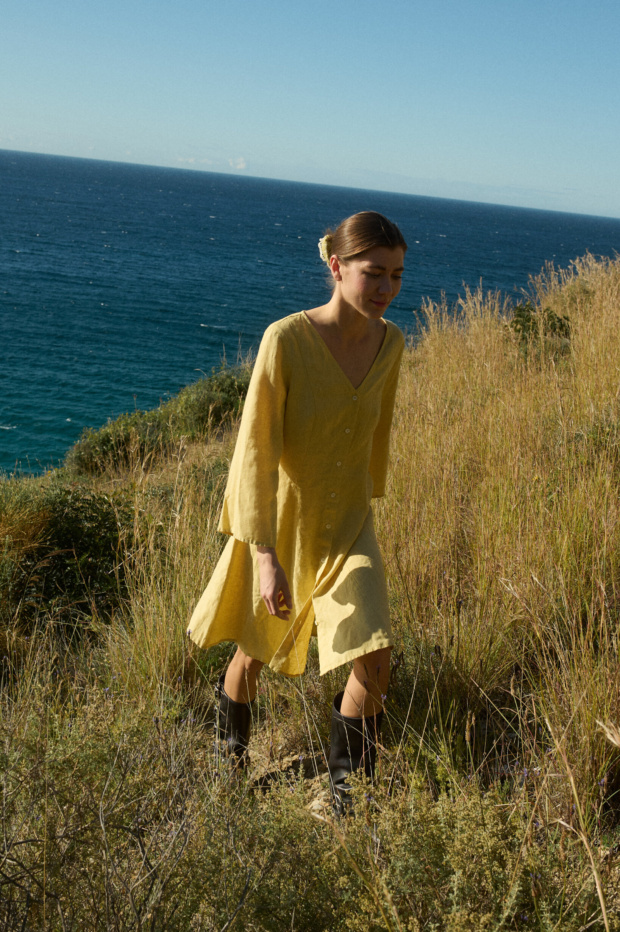 Model walking in a butter-colored linen dress featuring a V-neckline, button-down front, ¾ flared sleeves, and a relaxed A-line fit, with vertical linen panels and godet inserts for a flattering, flowing silhouette