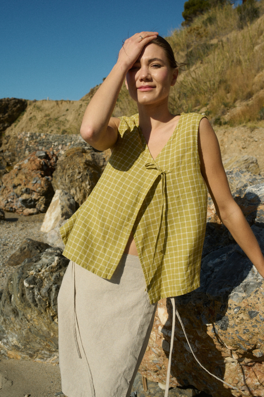 Model standing on rocky terrain, wearing a pickle checks linen sleeveless wrap top with a side tie, paired with a natural cotton blend skirt