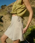 Model dressed in a sleeveless pickle checks linen top with a front tie, paired with a natural cotton blend wrap skirt that falls above the knee