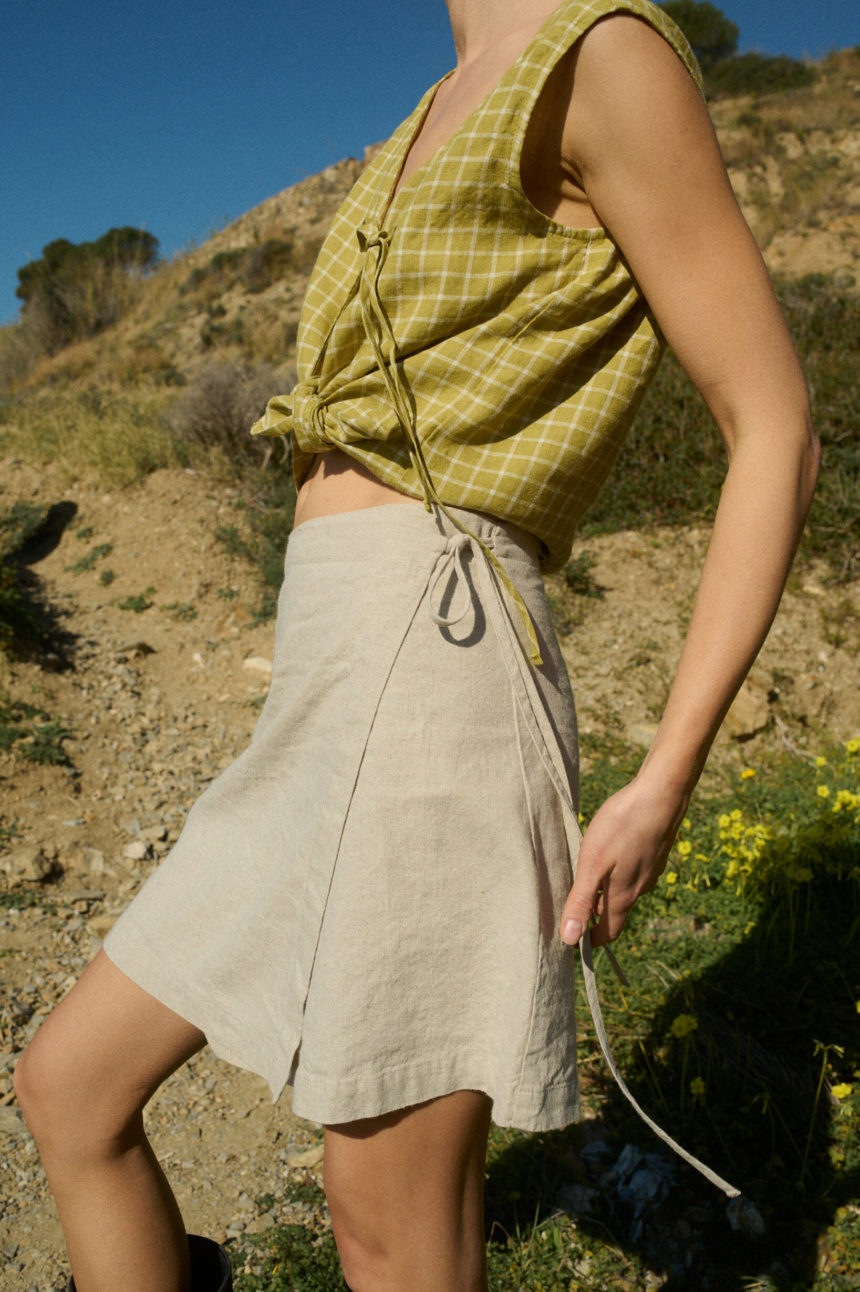 Model dressed in a sleeveless pickle checks linen top with a front tie, paired with a natural cotton blend wrap skirt that falls above the knee