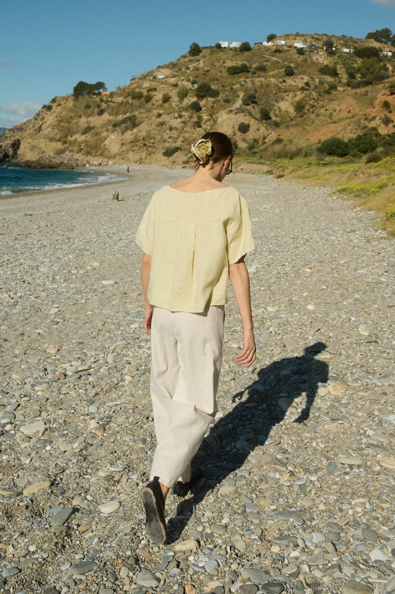 Model from the back wearing a pale yellow checkered linen blouse with a pleat in the back, paired with off-white linen-cotton blend wide-leg pants with an elastic waistband and patch side pockets.