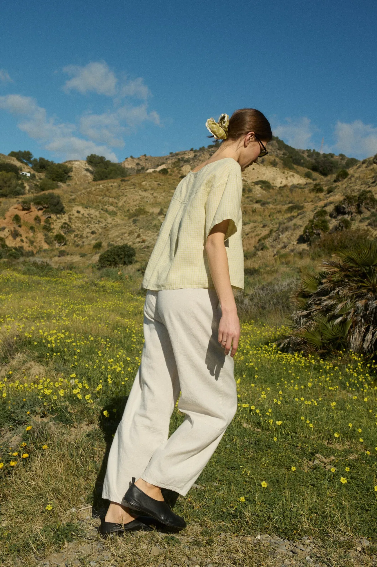 Model walking in a pale yellow checkered linen short-sleeve button-up top untucked, paired with off-white linen-cotton blend wide-leg pants with an elastic waistband and patch side pockets