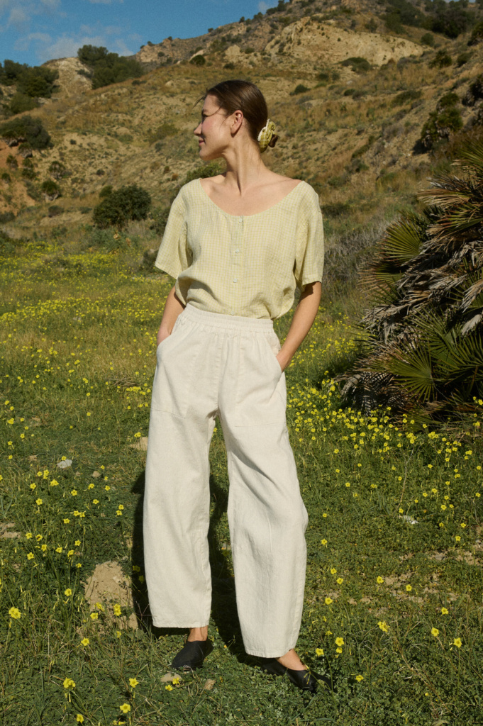 Woman wearing a flowing light yellow checkered linen button-up blouse with short sleeves and a scoop neck, tucked into high-waisted natural grey wide-leg pants