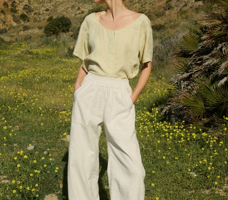 Woman wearing a flowing light yellow checkered linen button-up blouse with short sleeves and a scoop neck, tucked into high-waisted natural grey wide-leg pants