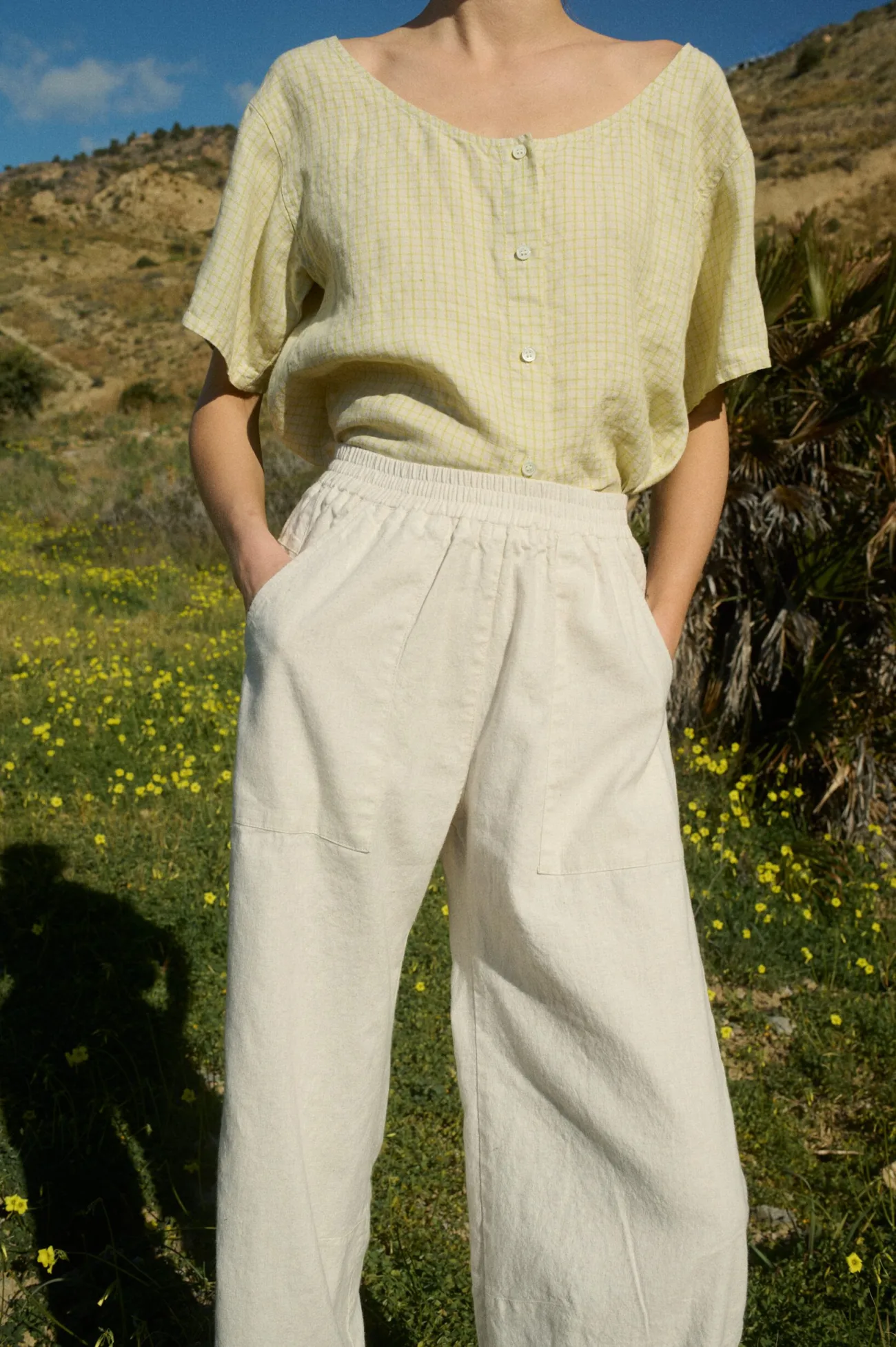 Model wearing a pale yellow checkered linen short-sleeve button-up with a scooped neckline, paired with off-white linen-cotton blend wide-leg pants with an elastic waistband and patch side pockets, standing with hands tucked into pockets