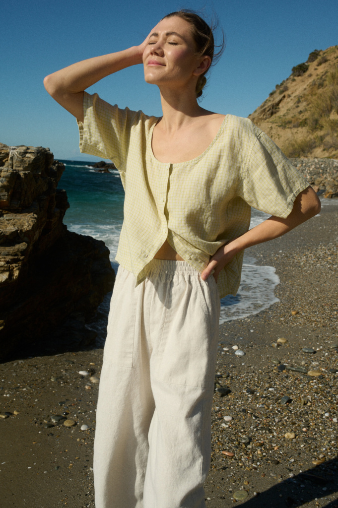 Model wearing a short-sleeved, button-up top in apple checks linen with a scoop neckline, slightly relaxed fit, and full-length buttons down the center front, paired with off-white wide-leg cotton pants with an elastic waistband