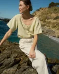 Model sitting on a rock, wearing a short-sleeved, button-up top in apple checks linen, tucked into off-white wide-leg cotton pants with an elastic waistband