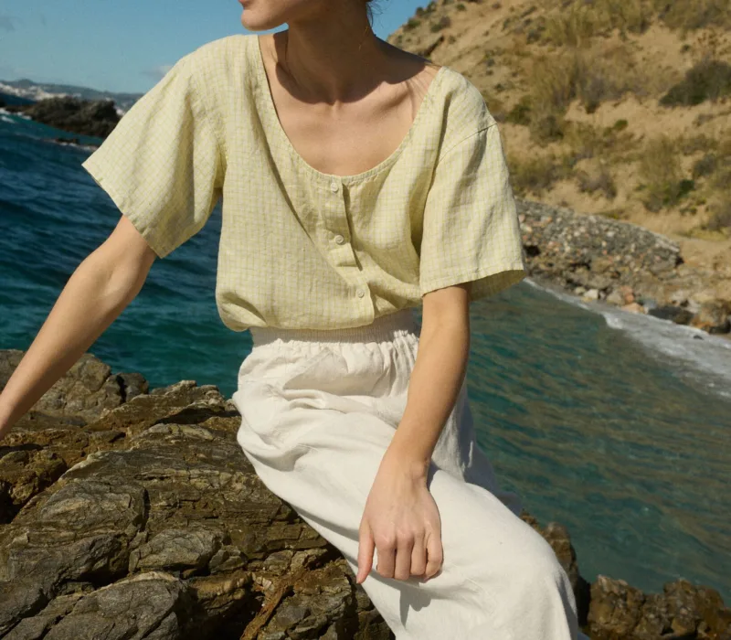 Model sitting on a rock, wearing a short-sleeved, button-up top in apple checks linen, tucked into off-white wide-leg cotton pants with an elastic waistband