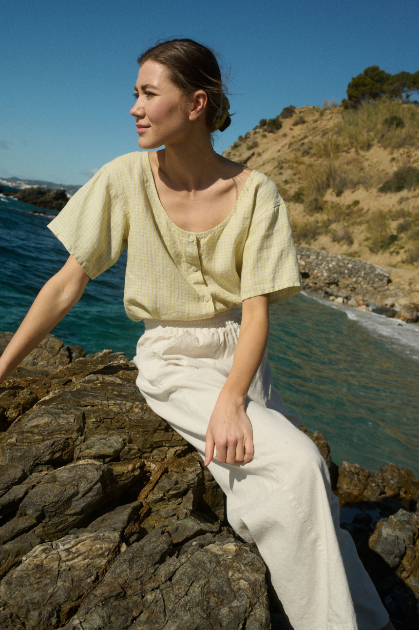 Model sitting on a rock, wearing a short-sleeved, button-up top in apple checks linen, tucked into off-white wide-leg cotton pants with an elastic waistband