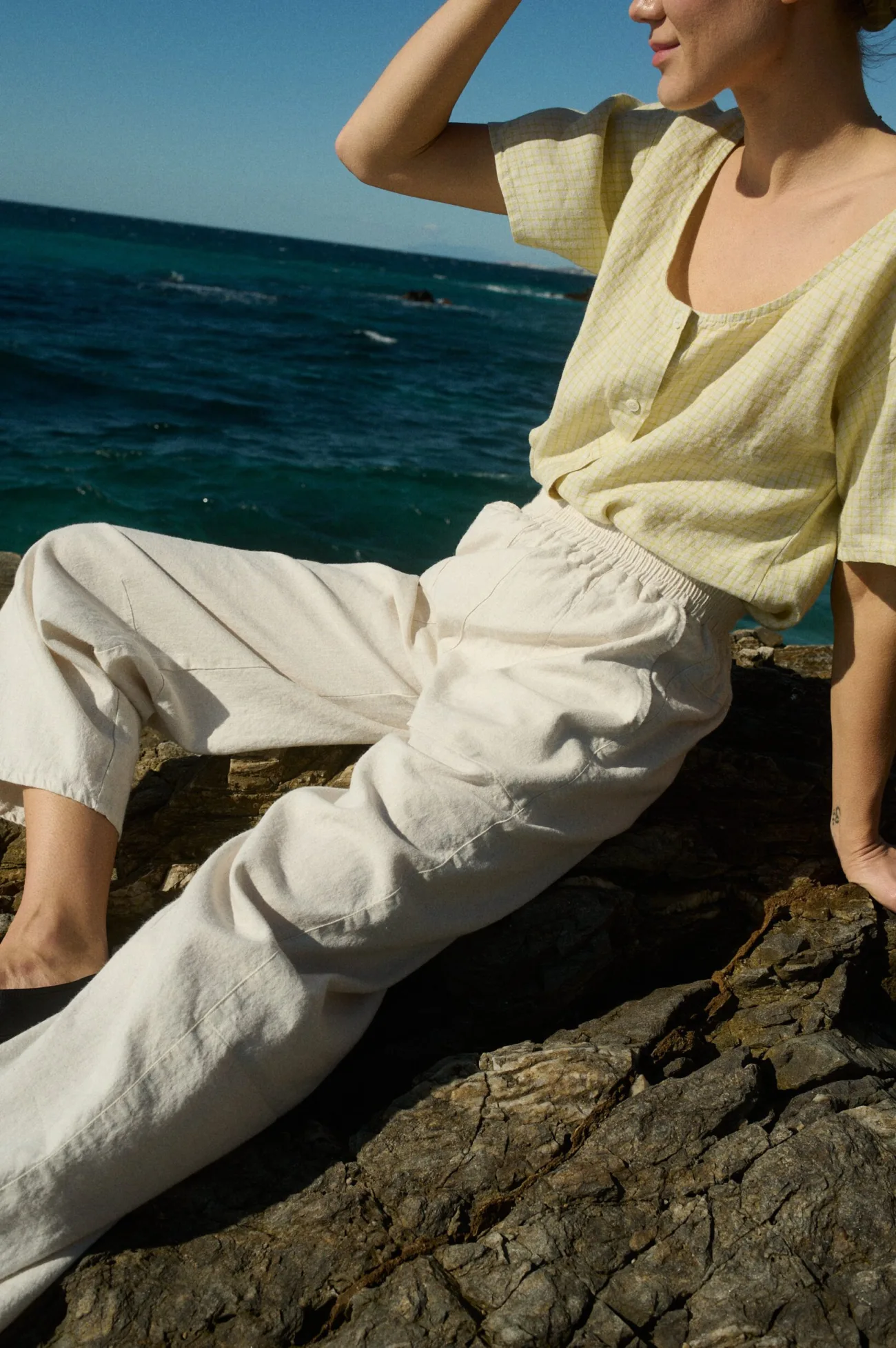 A model wearing a pale yellow checkered linen short-sleeve button-up with a scooped neckline, paired with off-white wide-leg pants made from a linen-cotton blend, featuring an elastic waistband and side pockets, set on a rock with the sea in the background