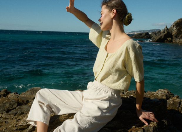 Model wearing a pale yellow checkered linen short-sleeve button-up shirt with a scooped neckline, paired with off-white wide-leg cotton blend pants with an elastic waistband and side pockets, sitting on a rock by the sea