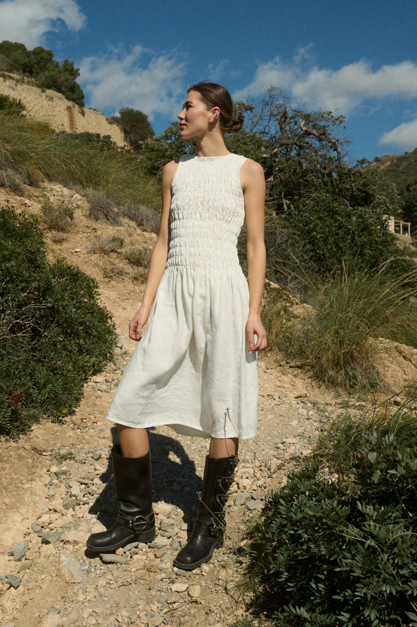 Woman wearing a knee-length white linen dress with intricate smocked detailing at the bodice, featuring a gathered waistline and a flowing skirt