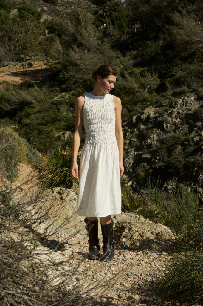 Model wearing a knee-length white linen dress with a relaxed A-line skirt, featuring intricate hand-smocked detailing across the bodice, a sleeveless design, and straight neckline