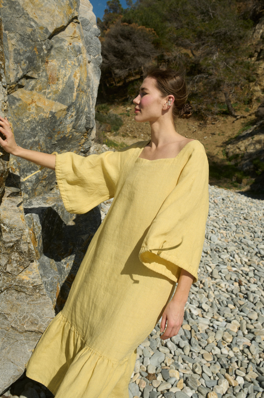 Model wearing a butter-colored midi linen dress with a wide squared neckline, ¾ length sleeves tied at the cuffs, and a softly ruffled hem for added movement