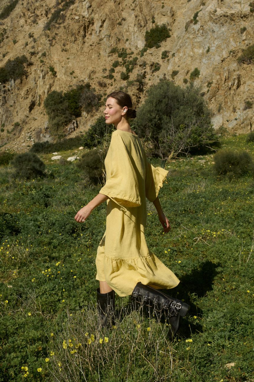Model walking in a butter-colored midi linen dress with a relaxed fit, featuring a squared neckline, ¾ sleeves with tie details, and a softly ruffled hem that moves with each step