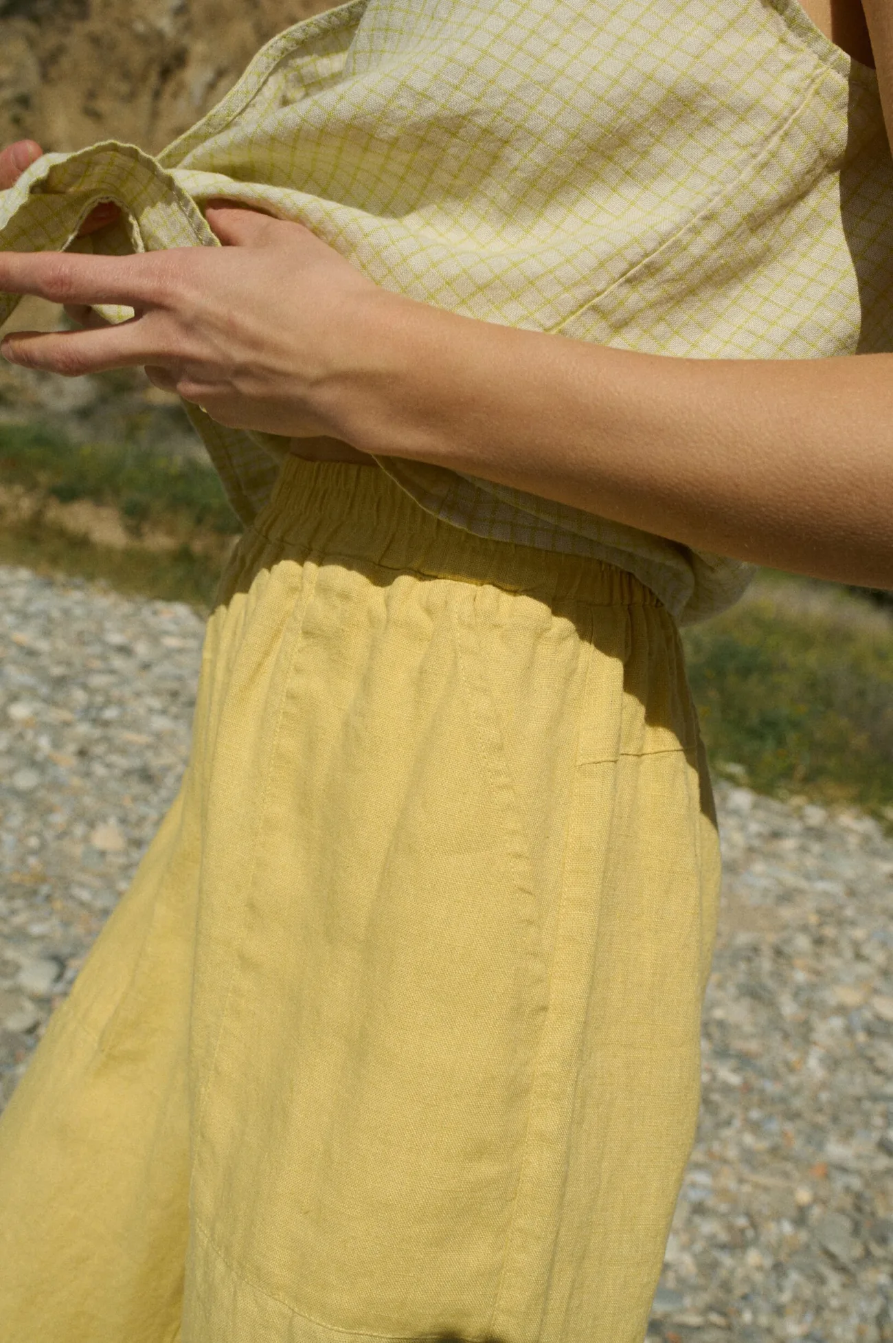 Close-up shot of a model highlighting the elasticated waist and side pockets of butter yellow wide-leg linen pants, paired with a pale yellow checkered sleeveless linen top