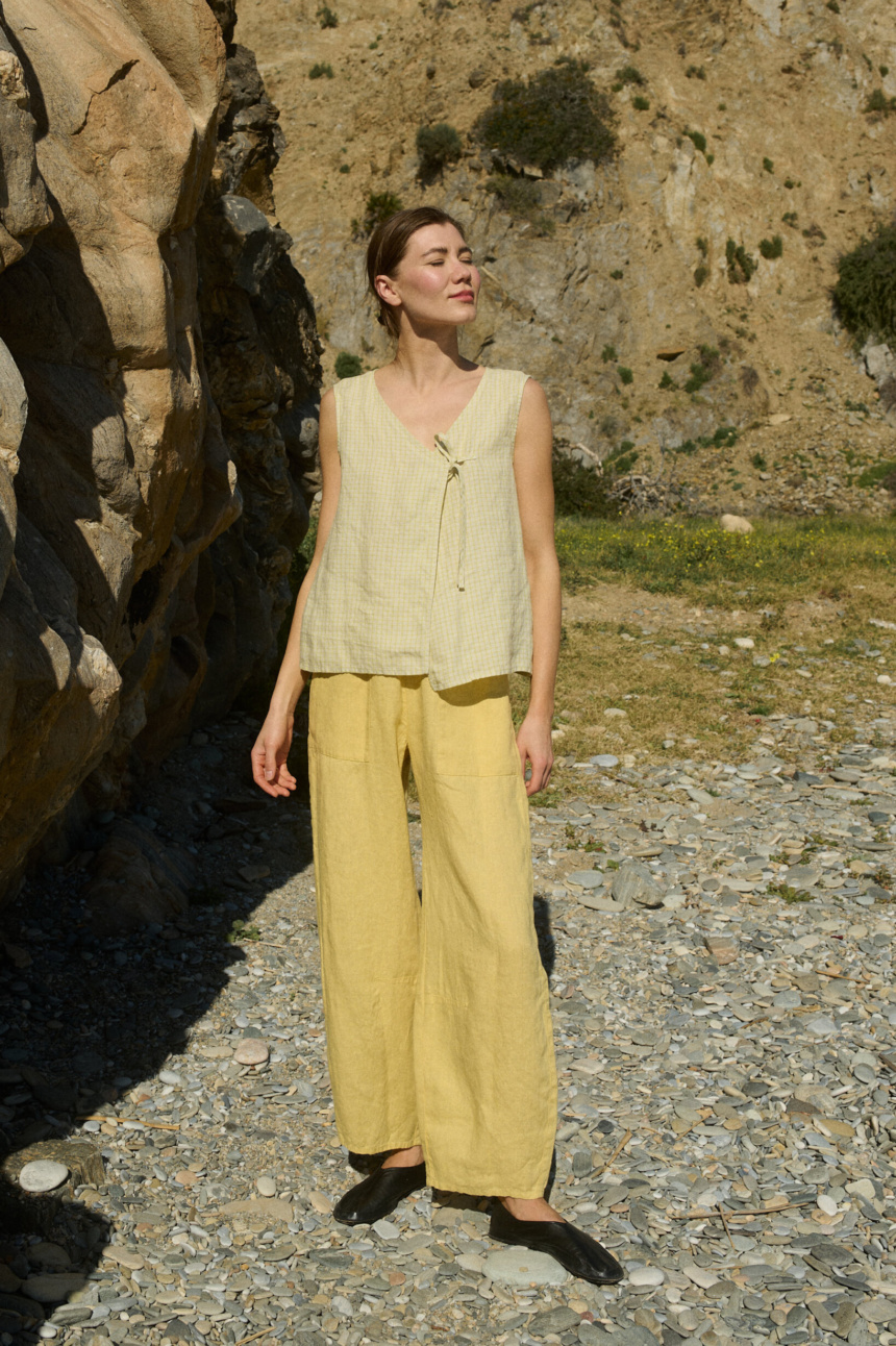 Model in a pale yellow checkered sleeveless linen top featuring a V-neckline, oversized fit, and a single side tie closure, paired with matching butter colored wide-leg pants for a breezy, effortless summer ensemble