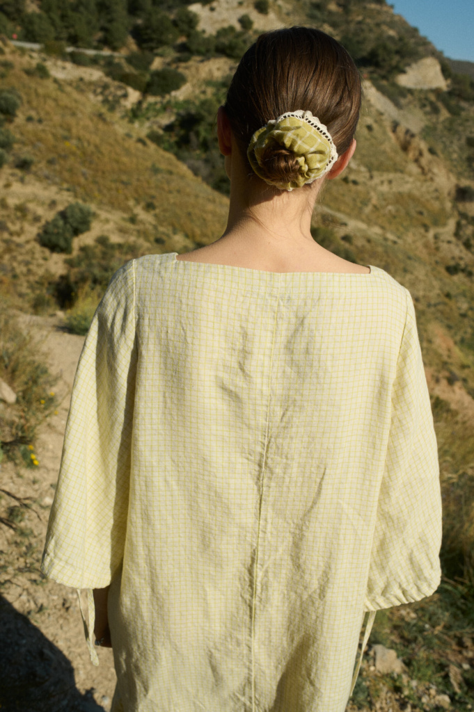 Back view of a model reveals a subtle apple checks pattern on the linen dress, with a centered vertical seam running down the back, wide relaxed sleeves, and a delicate linen hair scrunchie in pickle checks color adorning a neatly pulled-back bun