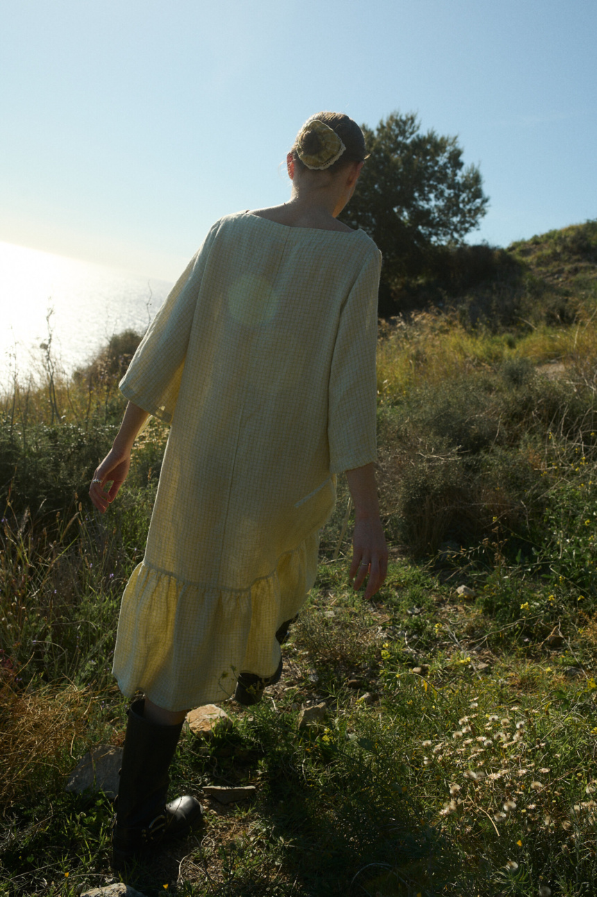 The back view showcases an apple checks linen dress with wide, relaxed sleeves, a centered vertical seam, and a charming linen hair scrunchie in pickle checks, adding a touch of flair to 1 neatly pulled-back bun