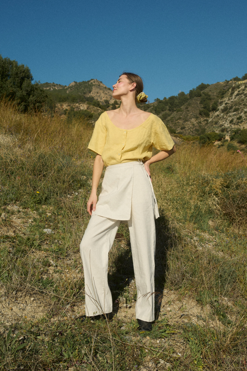 Model in nature wearing a bright yellow linen top and cream linen-cotton blend wide-leg pants with a wrap-around panel detail