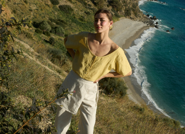 Model wearing a short-sleeved butter-yellow linen button-up top with a wide scoop neckline, tucked into flowing white cotton-blend wide-leg pants with a unique overlapping skirt panel
