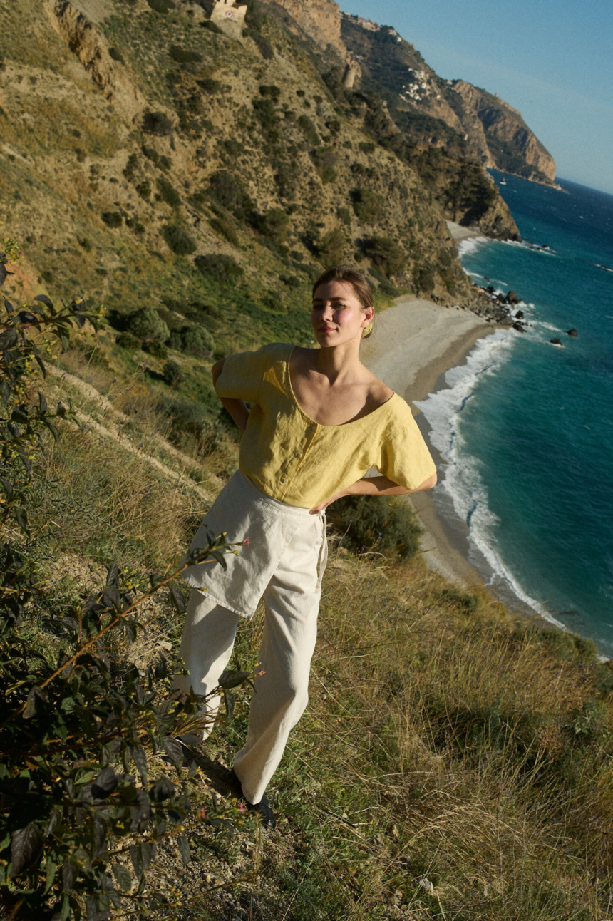 Model wearing a short-sleeved butter-yellow linen button-up top with a wide scoop neckline, tucked into flowing white cotton-blend wide-leg pants with a unique overlapping skirt panel