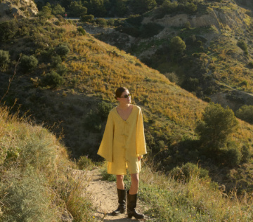 Model wearing a butter yellow linen tunic dress with wide, bell-shaped sleeves, a V-neckline, and a mid-thigh hem, paired with knee-high black leather boots and sunglasses, standing on a hiking trail with golden hills and evergreen trees in the background