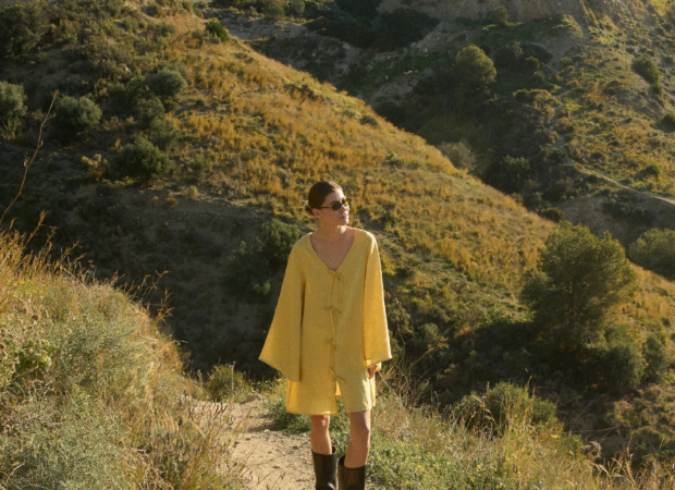 Model wearing a butter yellow linen tunic dress with wide, bell-shaped sleeves, a V-neckline, and a mid-thigh hem, paired with knee-high black leather boots and sunglasses, standing on a hiking trail with golden hills and evergreen trees in the background