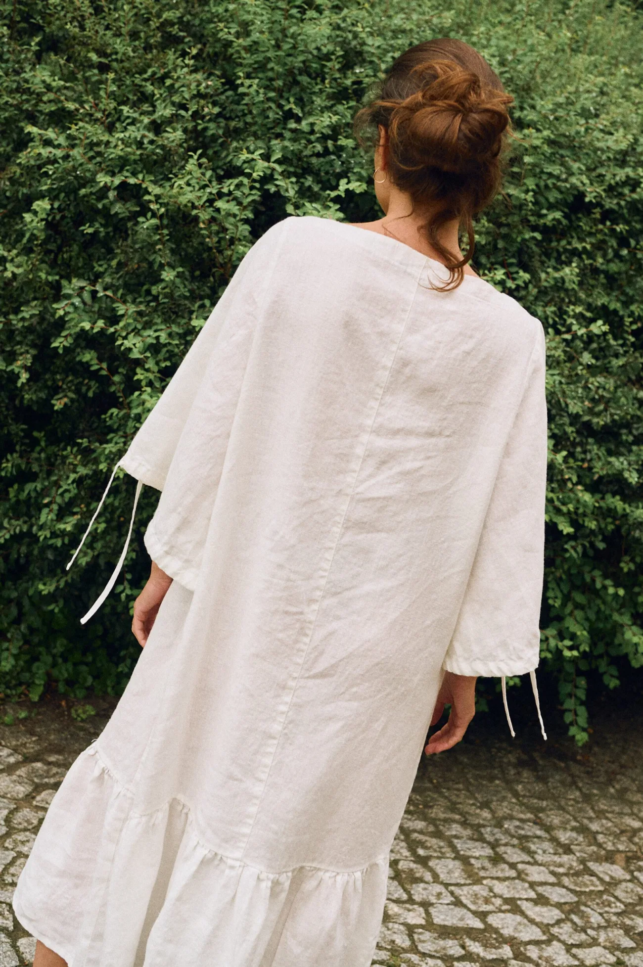 Back view of a model wearing an off-white linen midi dress with wide sleeves, adjustable ties, and a ruffled hem