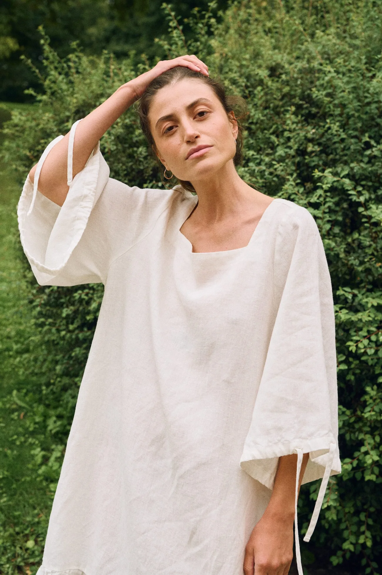 Model wearing an off-white linen dress with wide sleeves, adjustable ties, and a relaxed fit